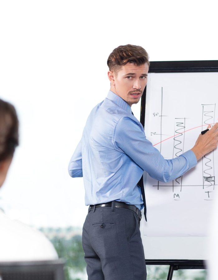 Young businessman standing at flipchart and pointing to graph and bar chart while turning back, looking and explaining ideas to audience seen partly